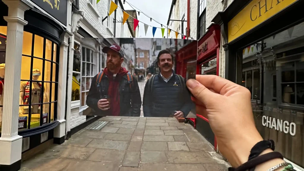 due uomini passeggiano per una strada con negozi. Uno ha una felpa rossa e un cappello da baseball, l'altro un piumino leggere. sono ted lasso e coach beard